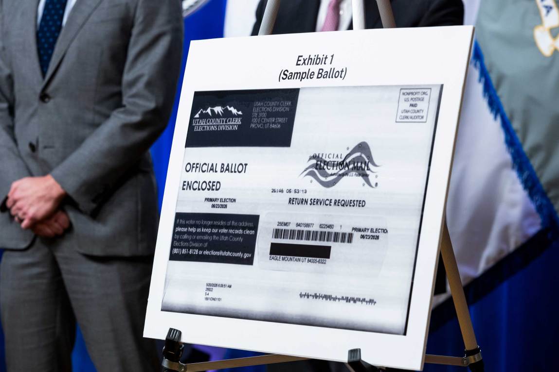 A poster labeled “Exhibit 1 (Sample Ballot)” displays a printed graphic of a mail-in ballot envelope from the Utah County Clerk Elections Division, set on an easel in front of officials in suits.