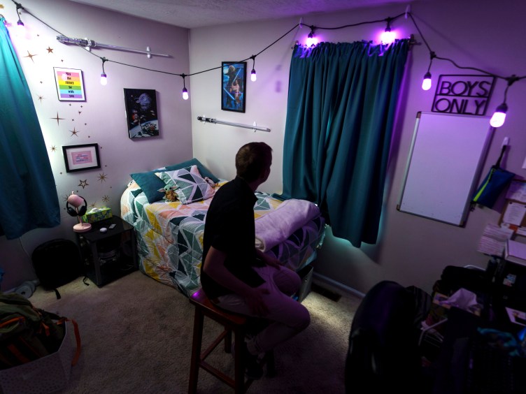 A man sits in a bedroom with his back to the camera. Decorations on the walls include Star Wars paraphernalia and a sign that says “boys only.”