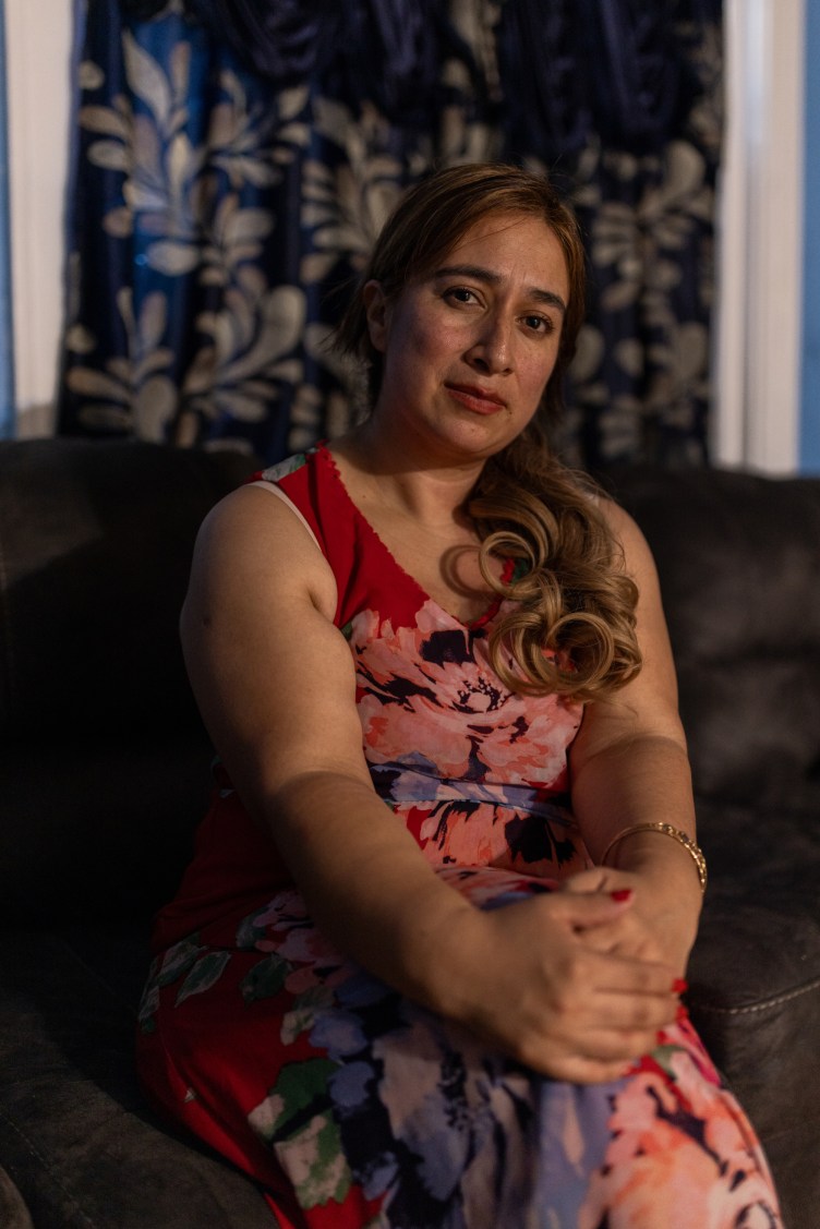 A woman sits in a dark room, looking at the camera, wearing a red floral print dress.