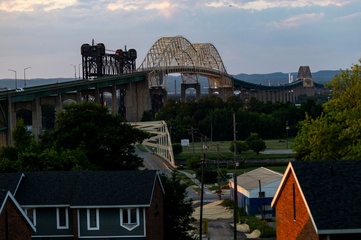 An elevated view overlooks residential house rooftops and lush green trees in the foreground. In the background is a massive bridge with three prominent yellow steel arches under a soft dusk sky.