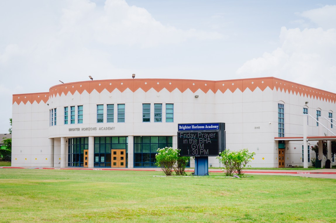 A two-story, light-beige Brighter Horizons Academy building features a curved front entryway and a distinct orange roofline decorated with a zigzag pattern. On the green lawn in front of the school, a digital sign announces “Friday Prayer in the BHA GYM 1:30 PM.”