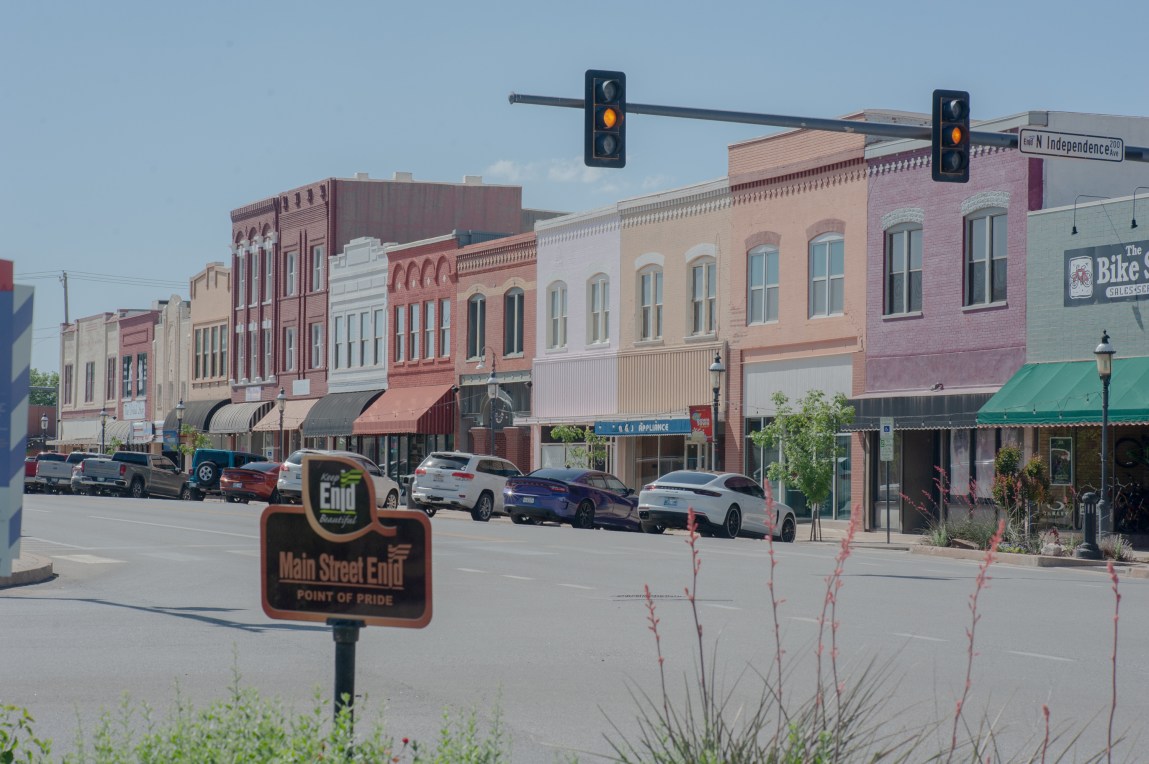 A sign reads, “Main Street Enid,” and behind it is the main street of a small town, with a row of buildings painted in pastel colors.