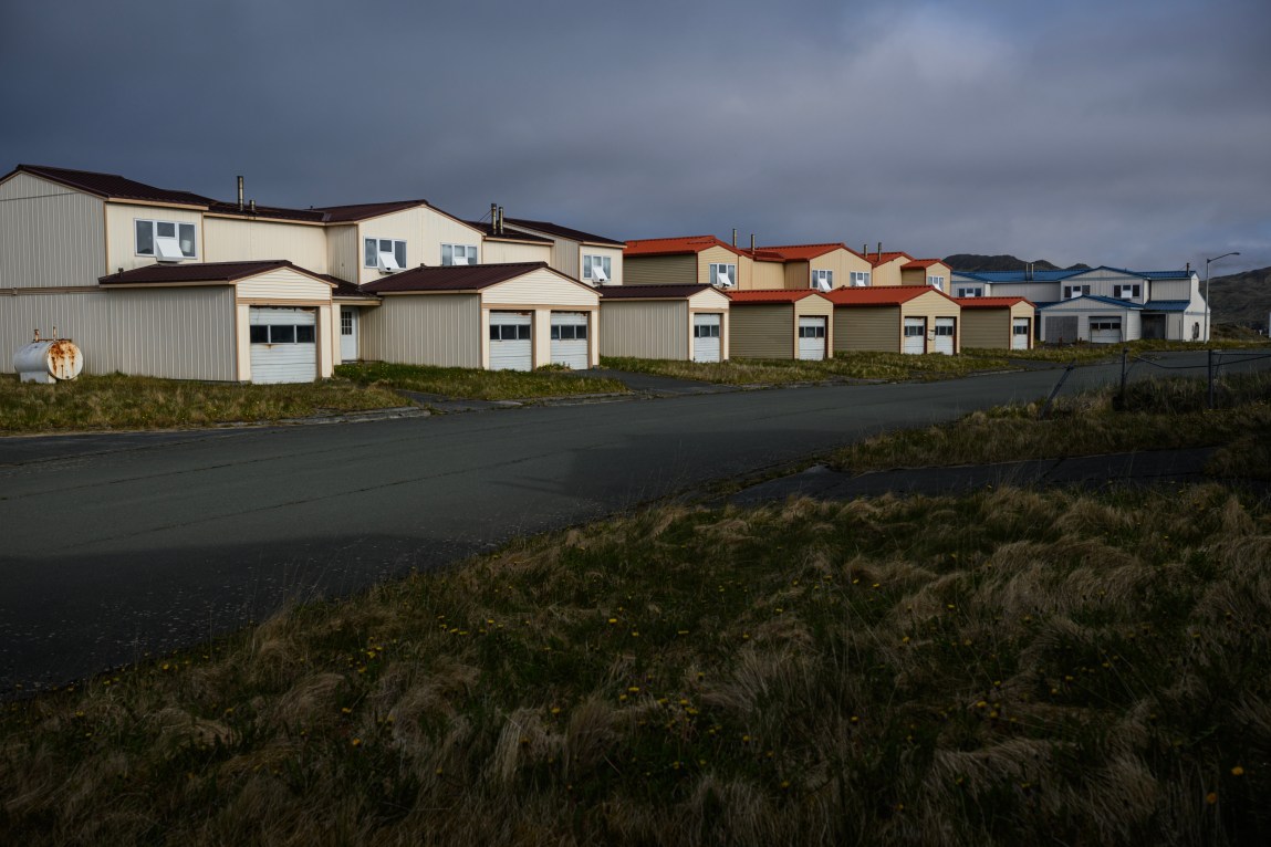 A row of houses sit on the edge of a paved road. An overgrown lawn is across the street. The sky is dark and cloudy.