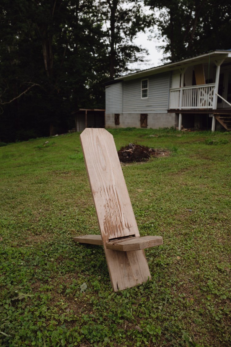 A simple, handmade wooden plank chair sits on a grassy lawn in front of a light blue house with a raised porch.