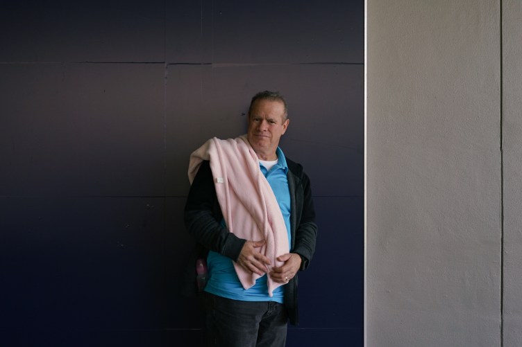 A man wearing a blue polo, black jacket and blue jeans, with a pink child’s blanket, stands against a wall.