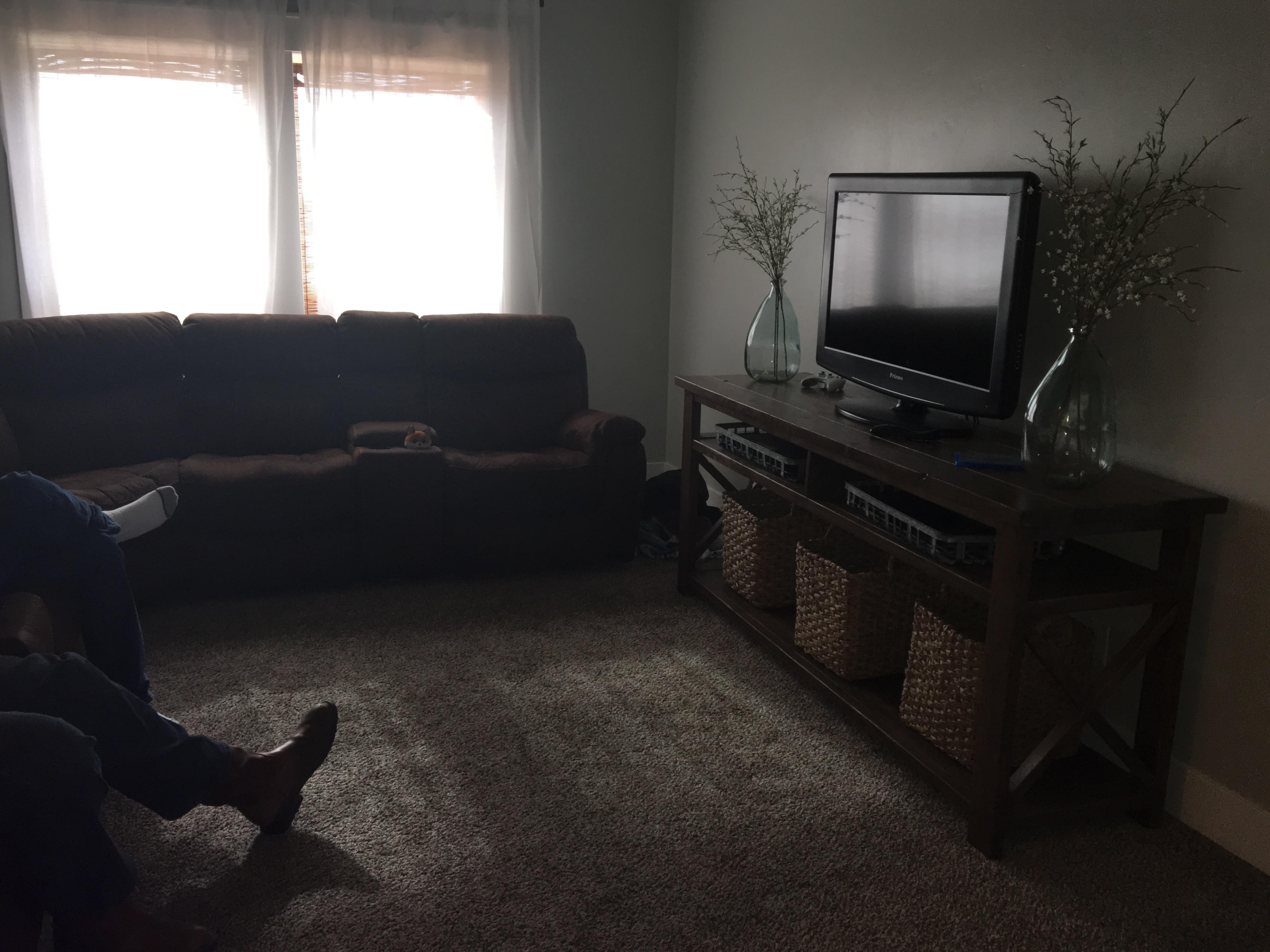 A dark, tidy room with two anonymous people crossing their legs while sitting on a couch. Only their feet can be seen: one clad in dress shoes, the other in a white sock. There’s a dresser with a TV, two glass vases and woven square baskets. Bright white light comes in through windows with white curtains.