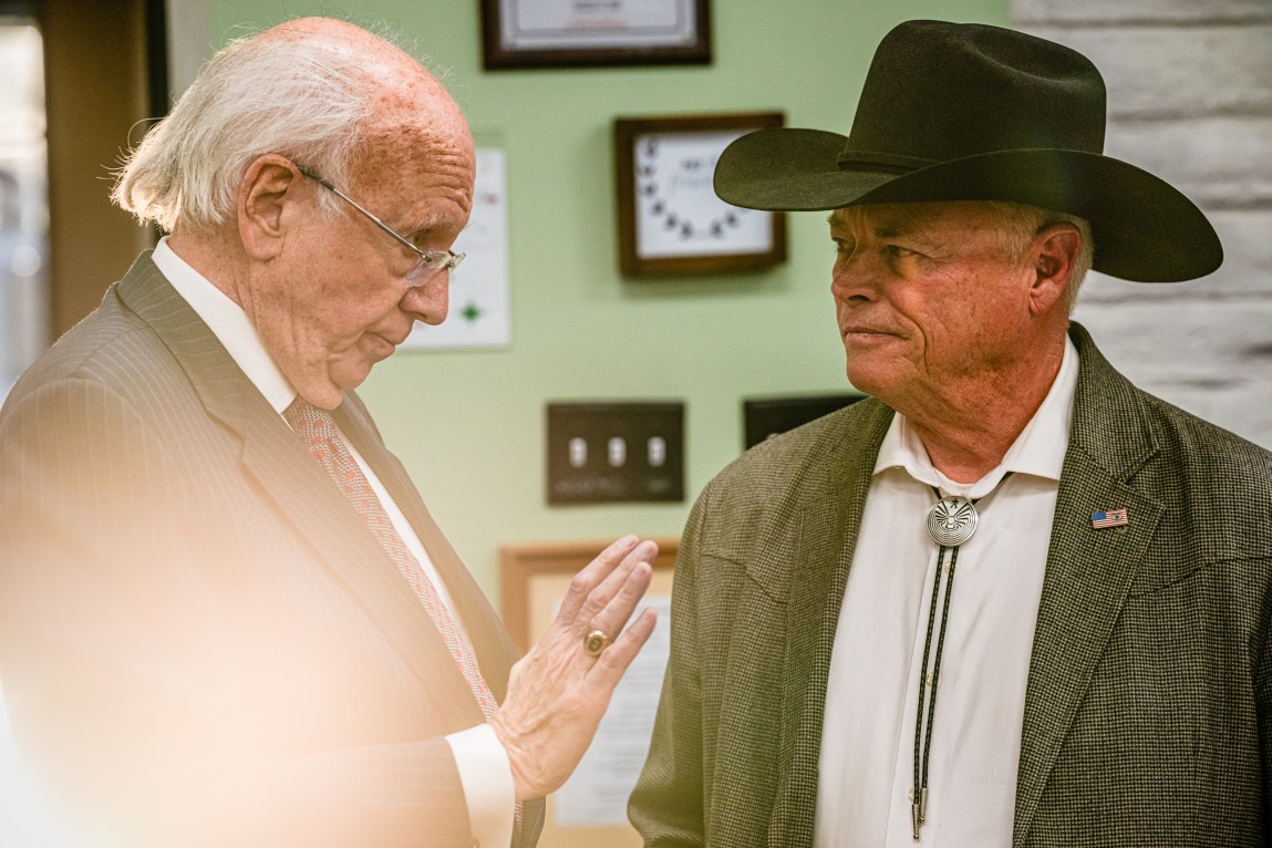 An older man in a coat and tie speaks with another man in a cowboy hat, suit jacket and bolo tie in the lobby of a community center.