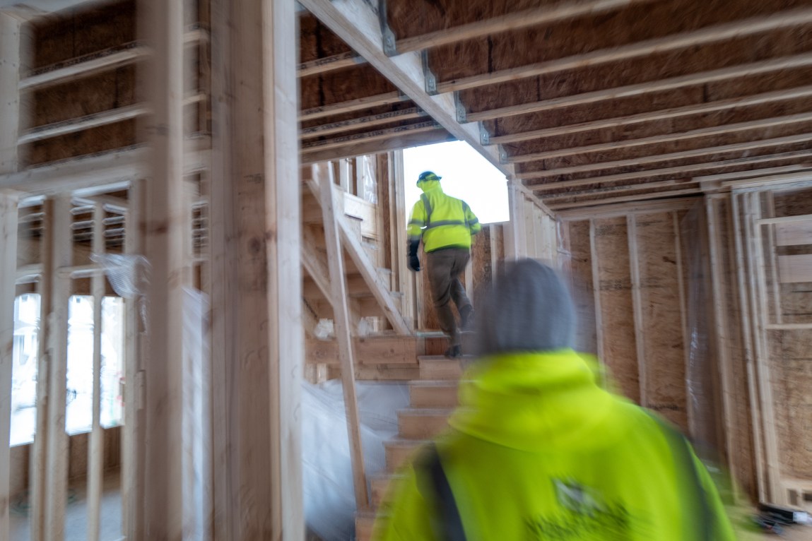 A blurry scene of two construction workers rushing through a partially built house, wearing bright high-visibility jackets.