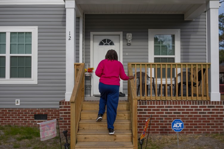 James walks up the wooden stairs built into a gray house with red bricks and white trim.