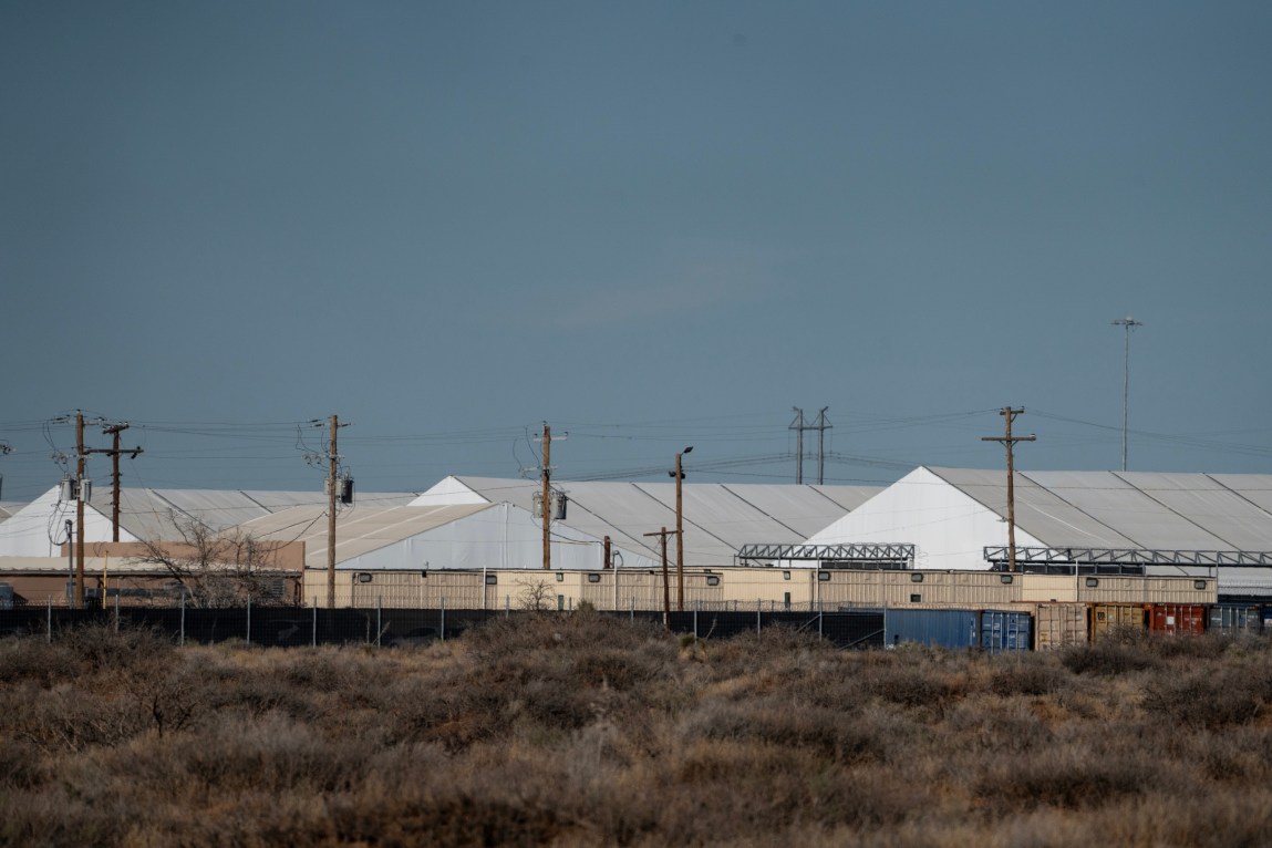 A distant, wide view of large white temporary tents and long beige structures surrounded by fencing and utility poles in a dry, desert landscape.