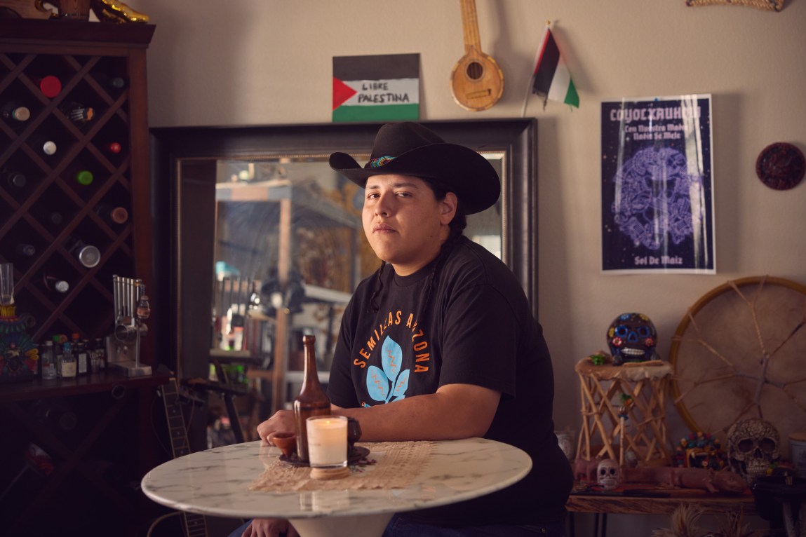 A young man wearing a black cowboy hat and black graphic T-shirt is seated at a small round table in a living room with a wine rack and sugar skulls behind him.