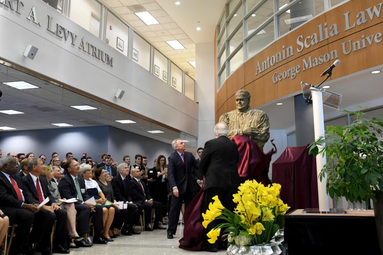 A crowd of people in business attire look on as two men pull a curtain down from a larger-than-life statue of Justice Antonin Scalia with his arms crossed.