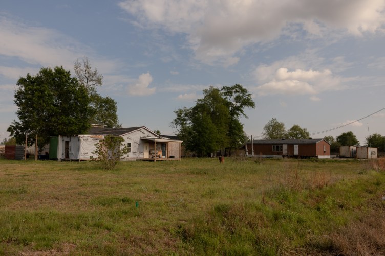 Three houses sit back in a large, grassy field with some short trees under a blue, cloudy sky.