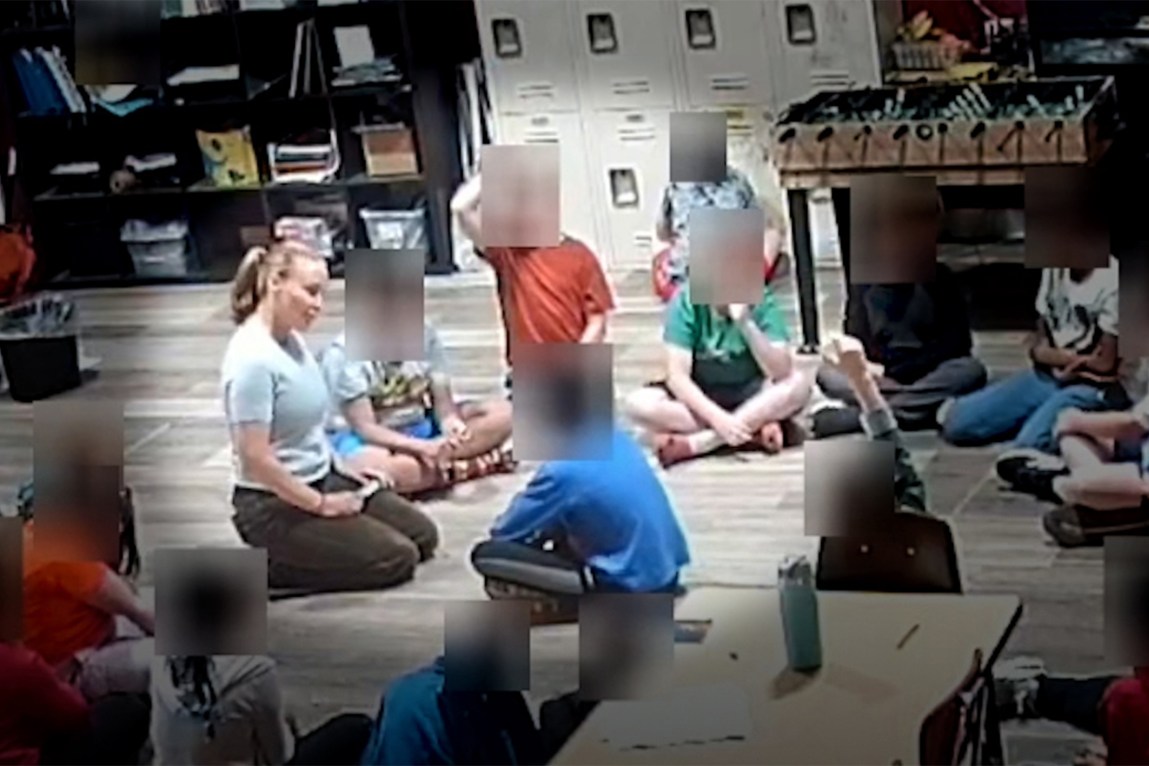 A circle of children sit on the floor in a classroom. One child sits cross-legged in the center of the circle, facing a kneeling woman wearing a ponytail and holding a cylinder in her hands. The children have blurred squares over their heads.