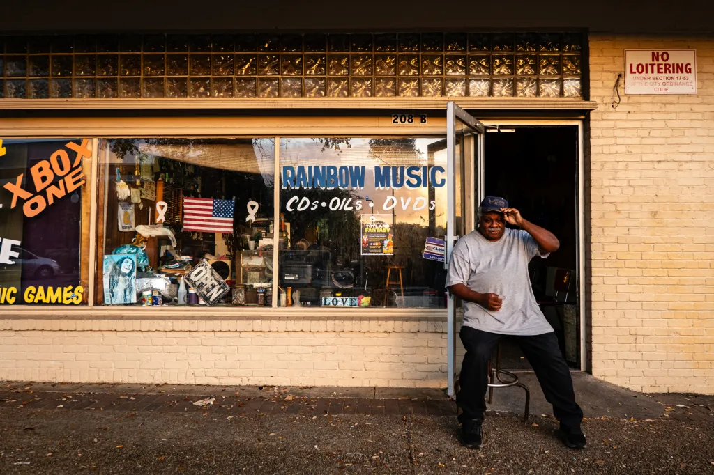 A man wearing a baseball cap sits outside a record shop. There is a warm light radiating on the storefront and the windows are full of audio equipment and bric-a-brac. One window reads, “Rainbow Music, CDs, OILs, DVDs”