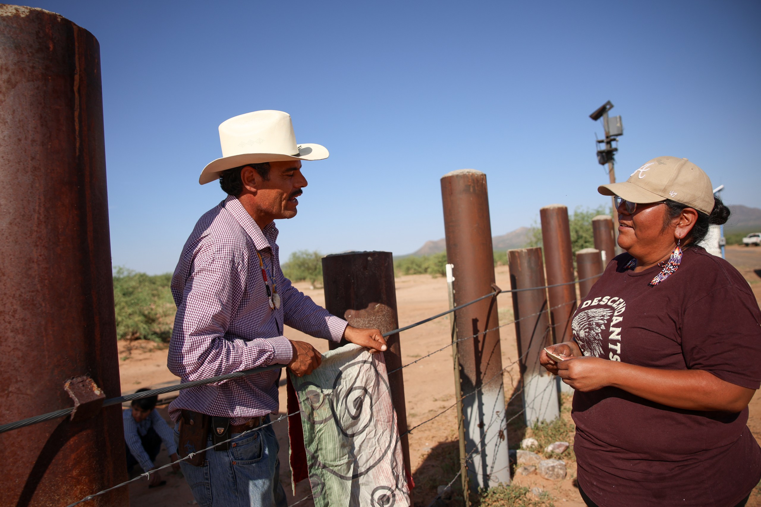 Two people visit across a fence made of barbed wire strung between thick metal posts in a desert landscape. In the background, a surveillance camera rises above them.