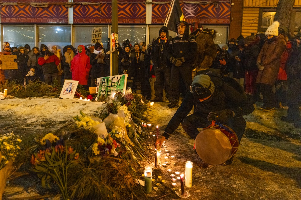 Flowers and candles piled in a semicircle on a ridge of snow. A spray-painted sign says “Alex Pretti,” and a crowd has gathered. One person holds a drum in one hand as they crouch and reach toward a candle with the other hand.
