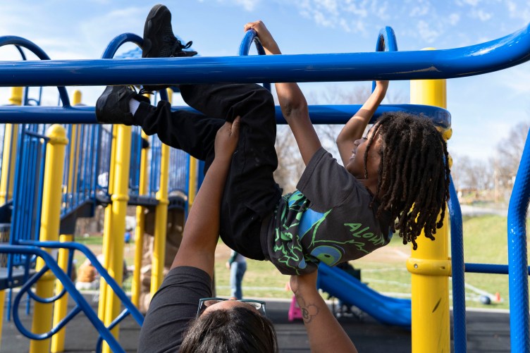 A woman lifts a smiling child up while he grabs the monkey bars and swings his feet up onto the play structure.