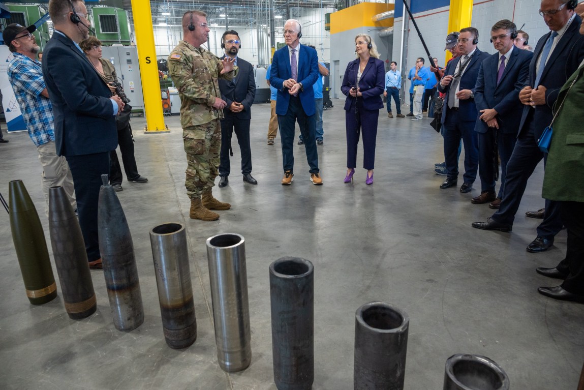 A man in an Army combat uniform speaks to a group of defense officials in a factory, in front of artillery shells at various stages of production.