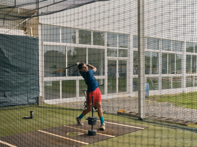 A baseball player inside a netted batting cage taking a swing at a ball off a batting tee. He wears a navy shirt, red shorts, high turquoise socks and white baseball gloves.