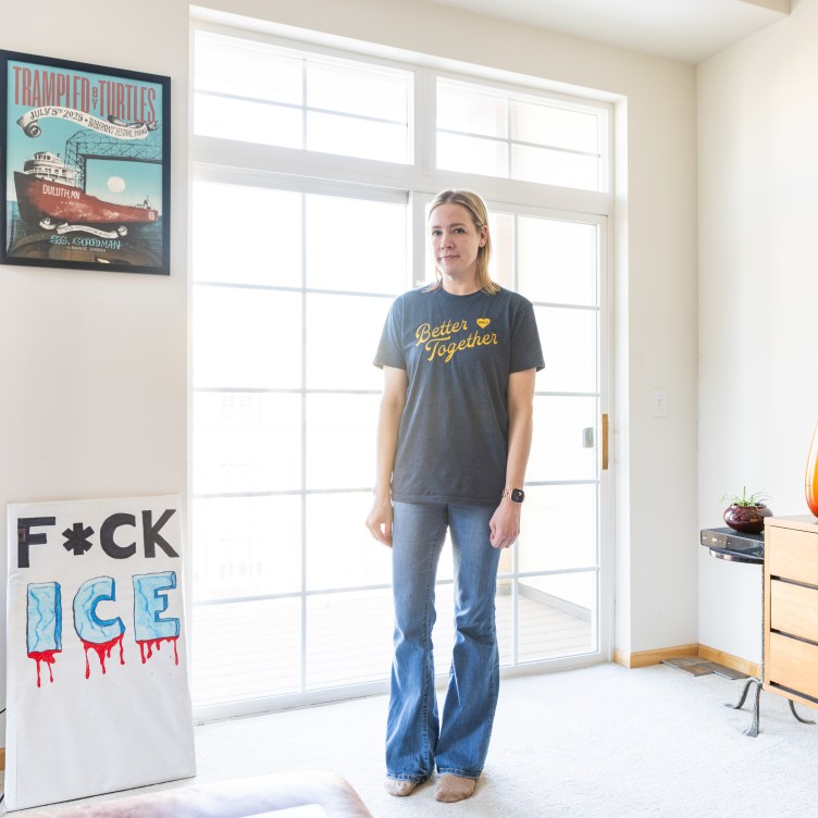 A woman stands in a bright condo, looking at the camera. A poster that says “Trampled by Turtles” is framed on the wall, and resting against the wall is a protest sign that reads “F*ck ICE.”