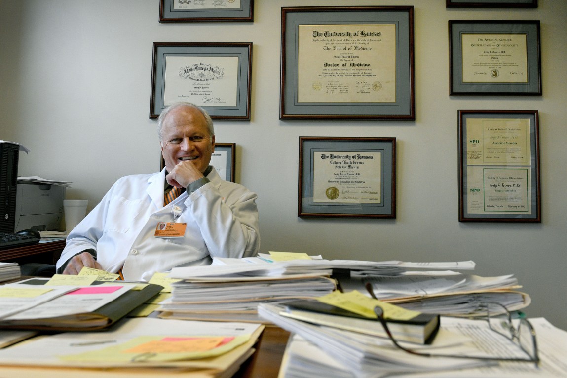 A smiling man with white hair wears a white doctor’s coat while sitting in front of a wall of framed certificates. The desk in front of him is piled with papers and a pair of glasses.