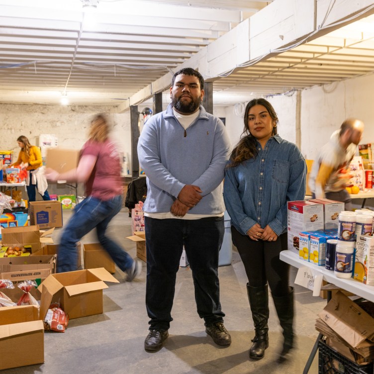 A man and woman stand together in a basement, looking at the camera. Around them are stacks of food, in cardboard boxes on the floor or on tables, and the blurred movement of several people moving and arranging the food.