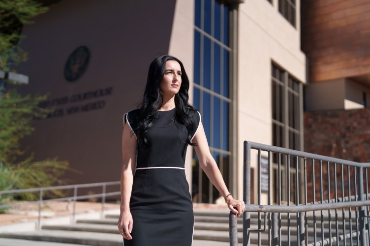 A woman with long black hair, wearing a black dress with white piping, stands while holding a banister in front of the U.S. district courthouse in Las Cruces.