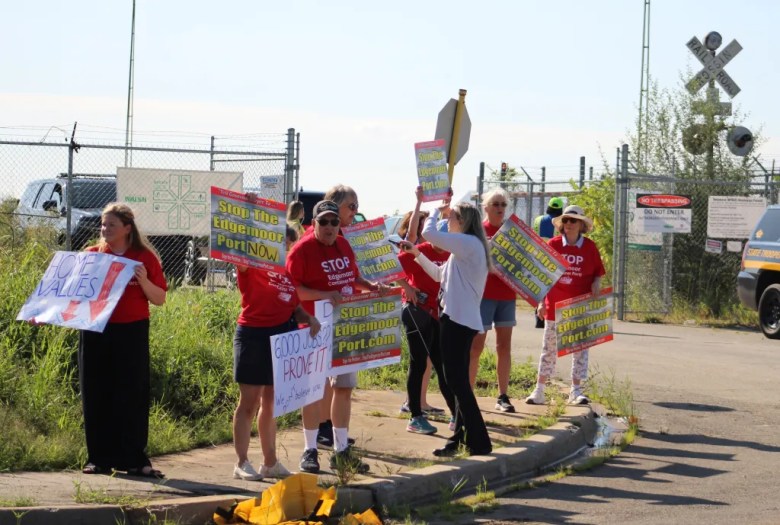 A group of protesters dressed in red shirts hold up signs protesting the Edgemoor port expansion.