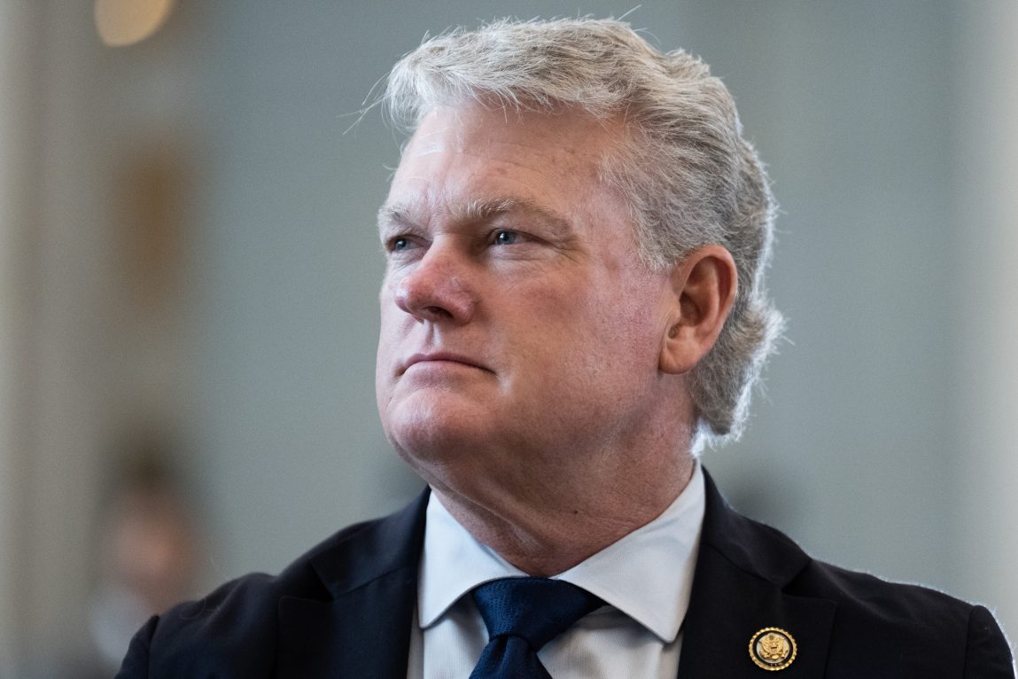 A man with gray hair wearing a suit and tie looks off camera to the left. The background is out of focus.