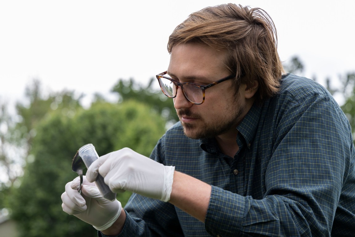 A man with brown hair and a beard wearing brown glasses, a teal long-sleeved button-down and white latex gloves holds a plastic test tube and a metal spoon. He’s staring intently at the spoon and test tube. Green trees appear out of focus in the background.