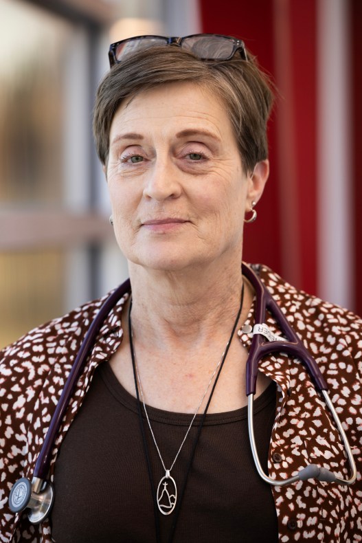 A woman with short hair, earrings, a stethoscope around her neck and glasses on top of her head looks directly at the camera.