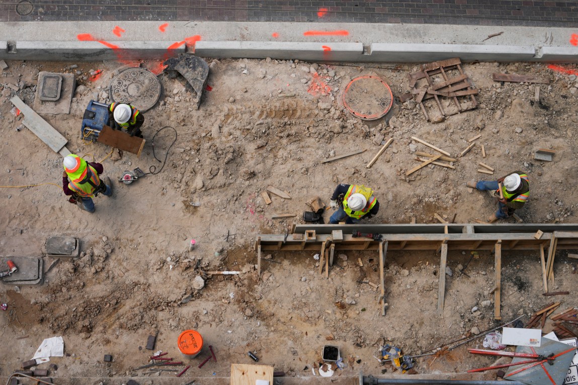 Four construction workers in yellow vests and white hard hats work on a large dirt path, as seen from above.