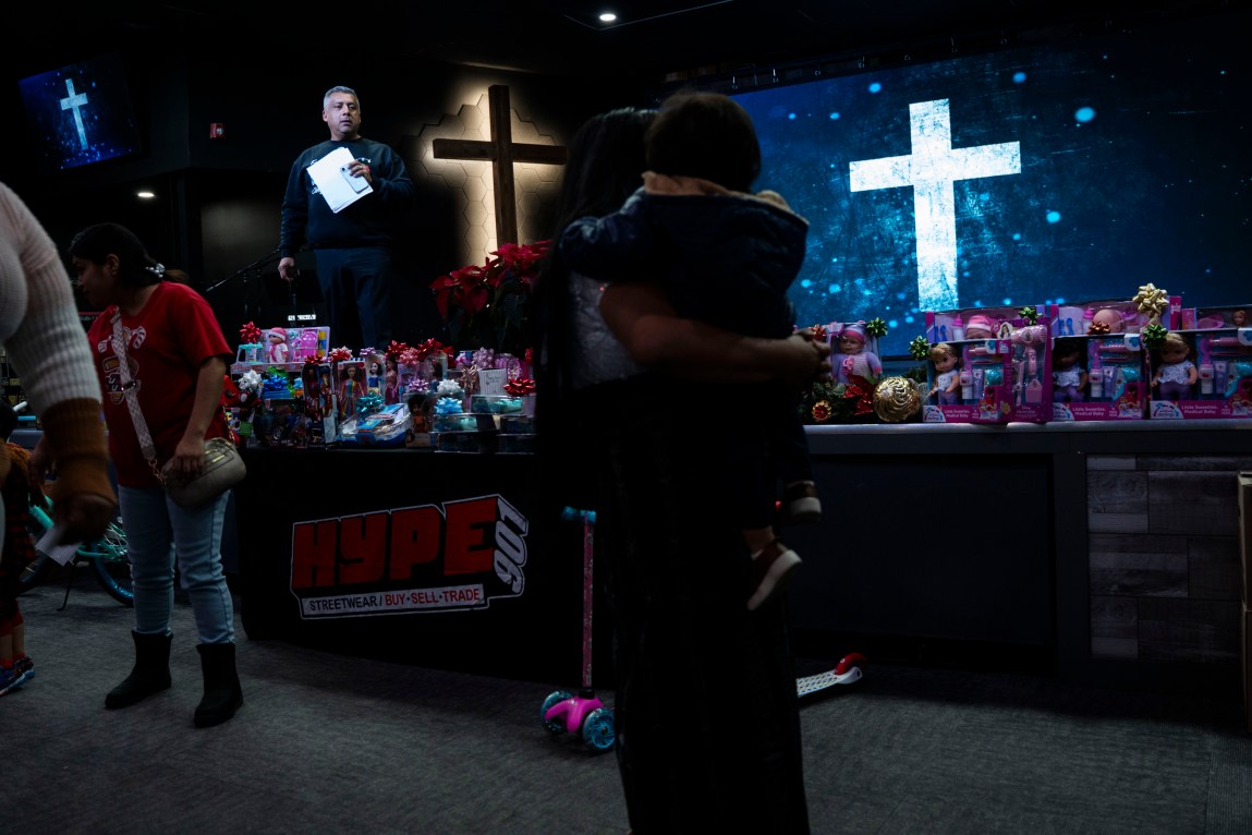 A man stands on a stage in a room decorated with several large crosses. Christmas gifts cover the stage. In the foreground, a person holds a child.