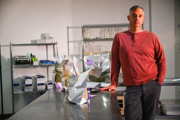 A man wearing a long-sleeve red shirt and gray pants leans against a metal table with green and yellow substances in bags. Metal shelves with equipment and glass containers sit against the wall behind him.