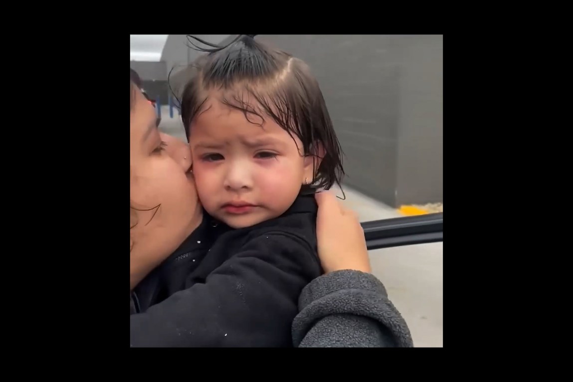 A close-up video still in which a 1-year-old girl with red, puffy eyes looks at the camera while her mother kisses her on the cheek.
