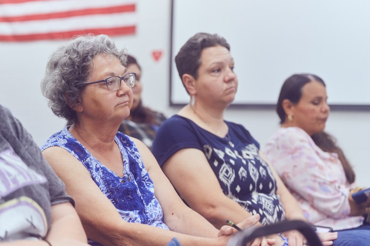Three women seated in an audience in a white room with a whiteboard and an American flag on the wall behind them.