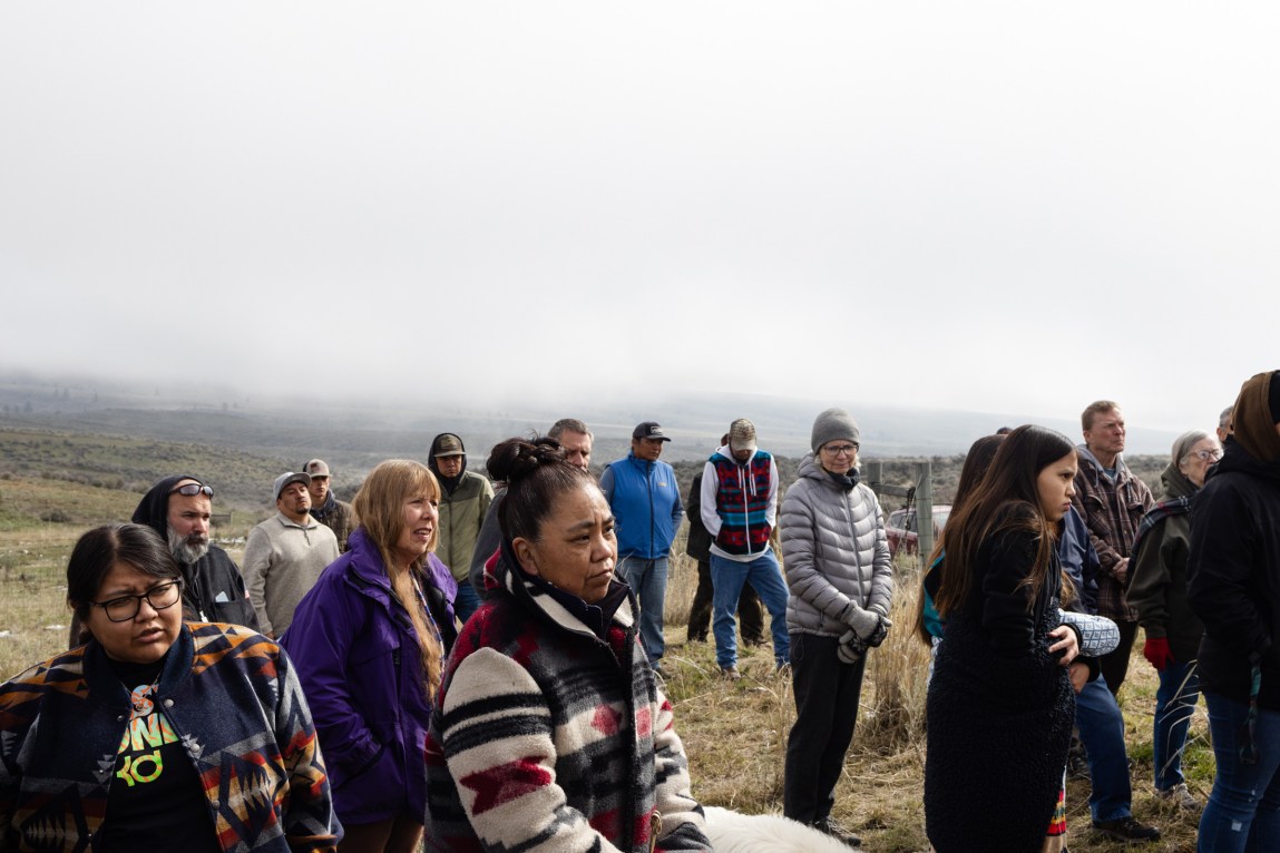 A group of people wearing jackets stand on a grassy mountain with white fog in the background.