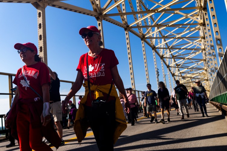 Two women wearing matching red Canada-themed shirts and red caps walk across a bridge amid a crowd of pedestrians. The sunlit bridge frame towers overhead against a clear blue sky.