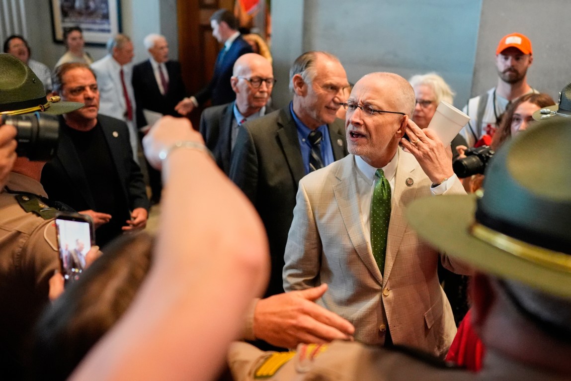A man in a suit holds his finger to his ear in an “I can’t hear you” motion as he walks through a government building. In the foreground are a crowd of people raising their arms at him, cameras pointing at him and people in uniform holding the crowd back.