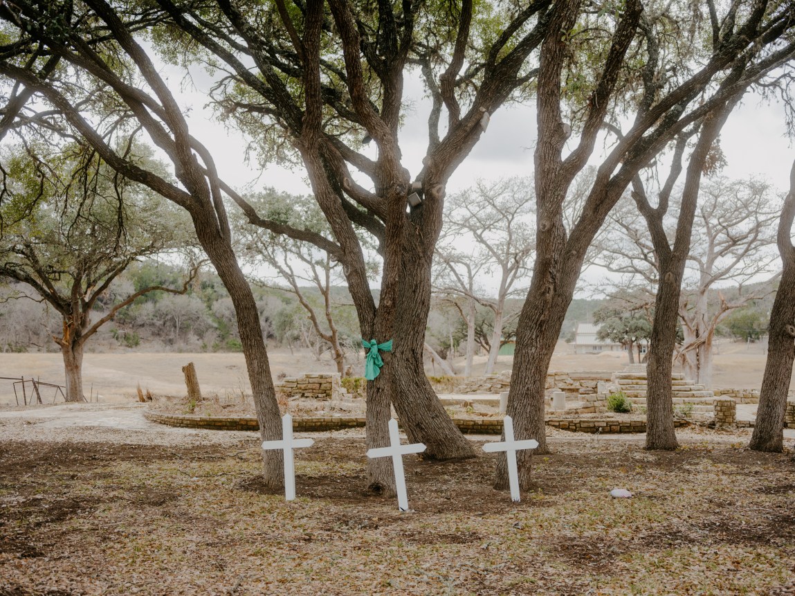 Three white crosses stand in the ground under a canopy of trees near a river on a cloudy day.