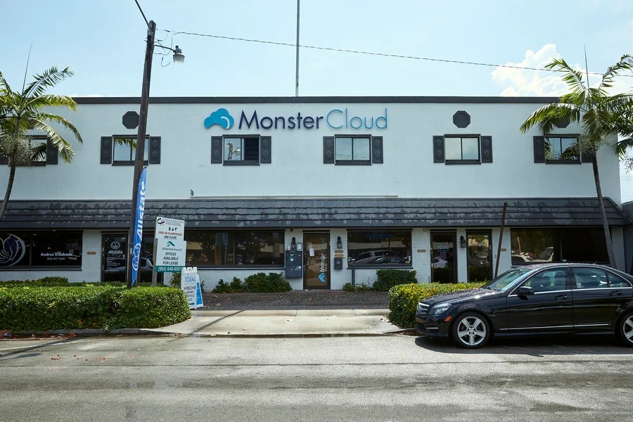 Palm trees and green hedges grow in front of a white two-story building with a sign that reads MonsterCloud.