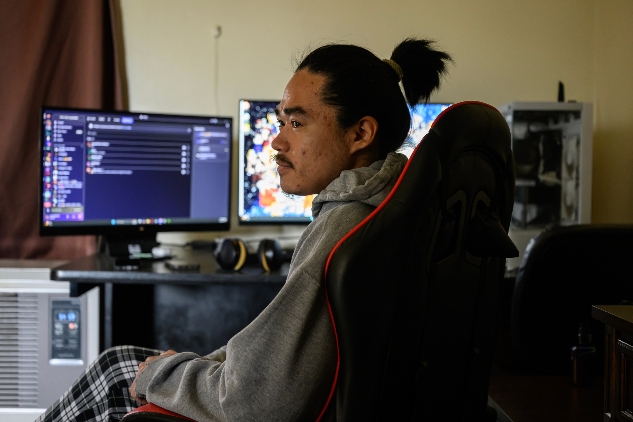 A man with a ponytail and moustache wears plaid pants and a gray hooded sweatshirt. He sits in a gamer chair in front of multiple computer monitors and stares away from the camera to the left.