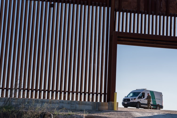 A man walks in front of a U.S. Border Patrol van, parked near a large entrance in a tall section of the steel bollard U.S. border wall with Mexico.