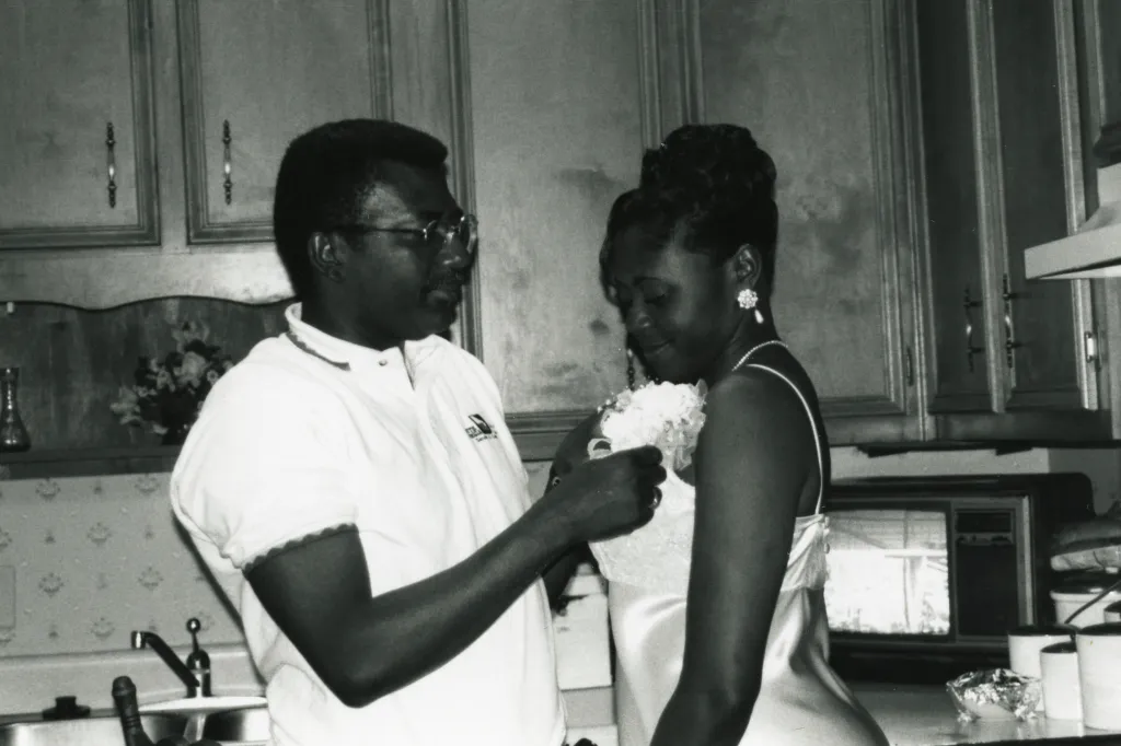 A man and young woman stand in a kitchen. The man is attaching a corsage to the woman’s dress. I