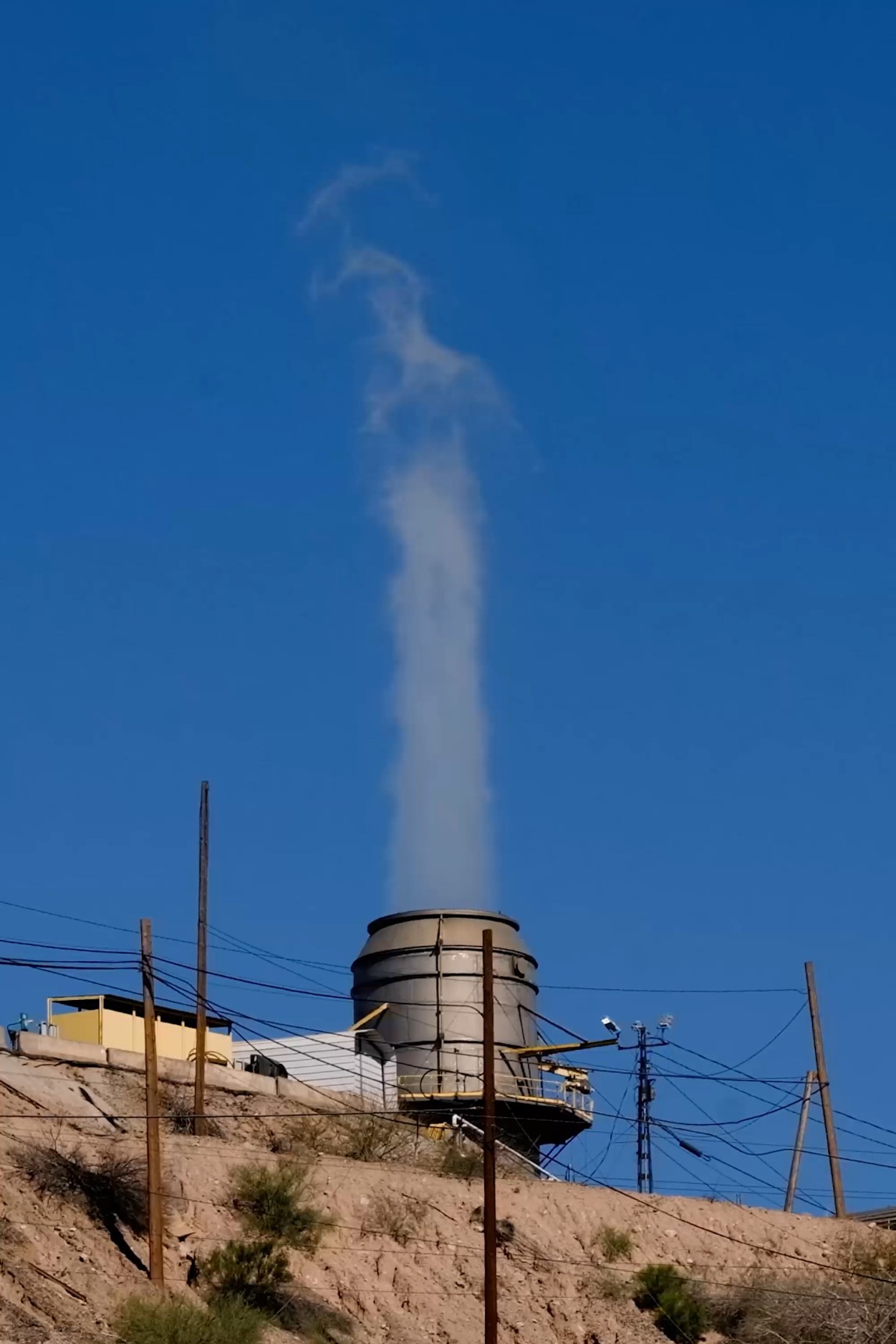 An industrial cooling tower sits on a dry, rocky incline against a clear blue sky, emitting a steady, vertical plume of steam.