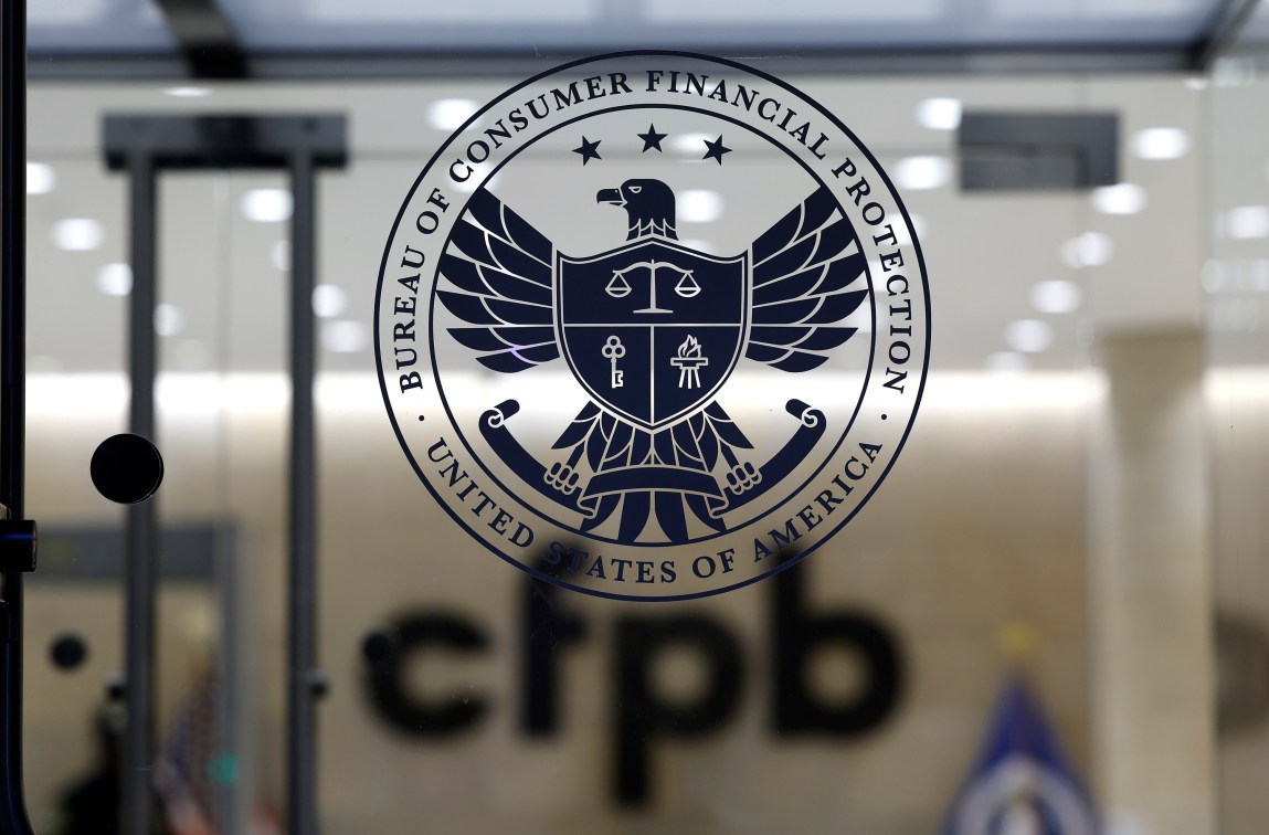 The CFPB logo on glass. Out of focus behind the logo, “CFPB” appears on a wall near a metal detector and a flag. Rows of circular lights line the ceiling.