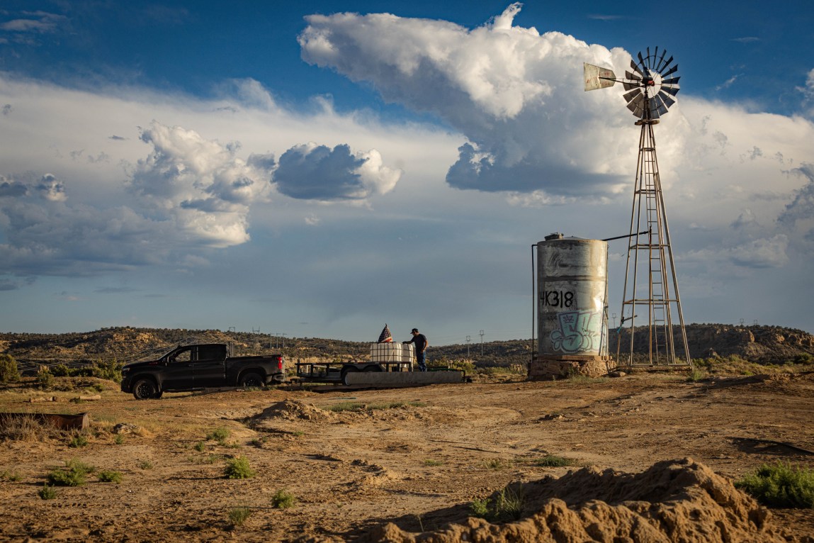 A dark pickup truck is parked near a tall, silver water tank and a metal windmill on a dry, dirt lot under a dynamic sky filled with fluffy white clouds.