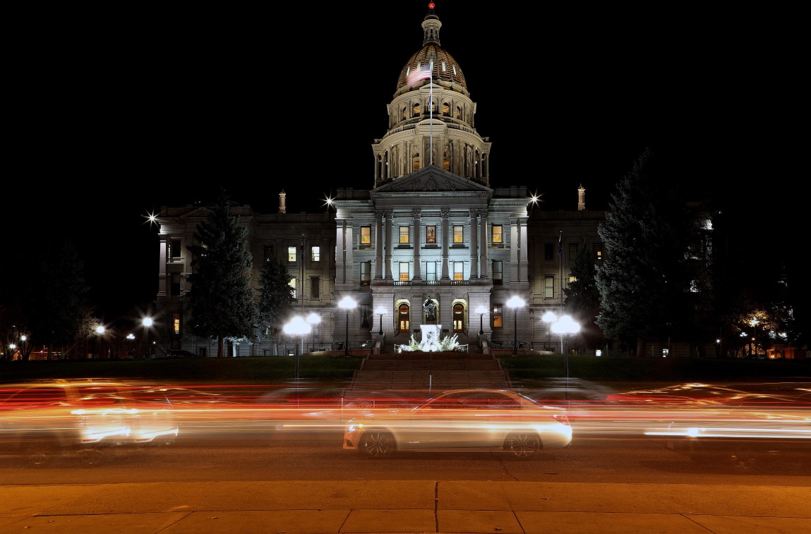 The Colorado State Capitol lit up at night. In the foreground, cars speed by as their headlights and taillights form long streaks of color.