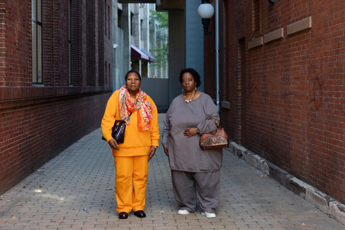 Two women stand in an alleyway flanked by brick buildings. One woman wears a scarf, orange pants and an orange shirt; the other woman wears a gray shirt and pants, along with jewelry and a brown purse.